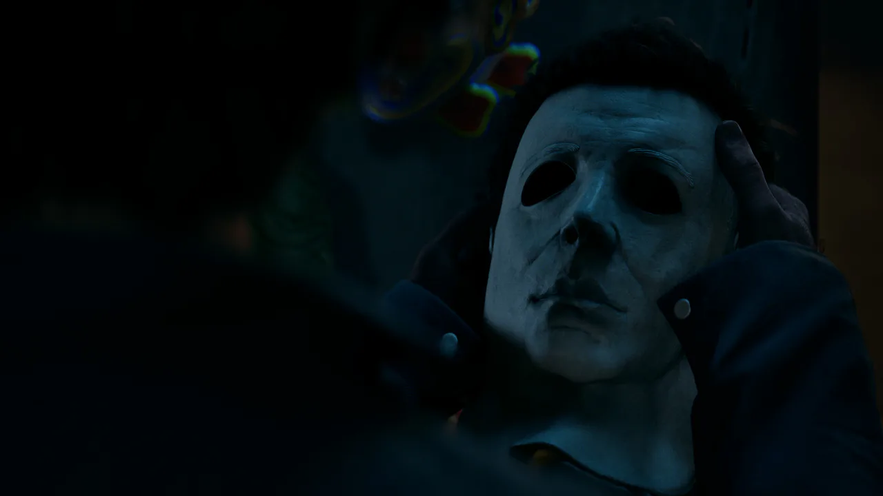 Primer plano de la icónica máscara de Michael Myers siendo sostenida por manos humanas en una atmósfera oscura del videojuego Halloween: The Game.
