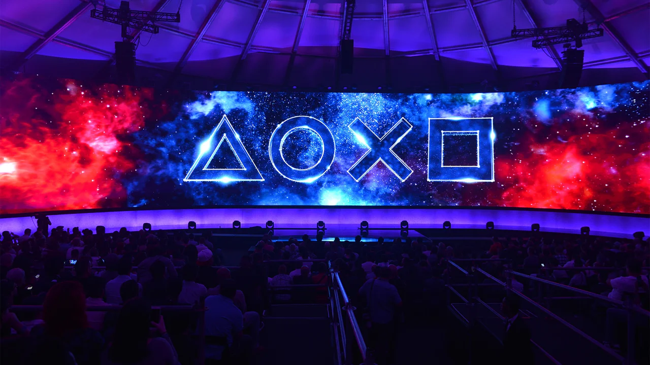 Vista panorámica de un escenario de evento de PlayStation, donde una gran pantalla curva exhibe los icónicos símbolos de la marca (triángulo, círculo, cruz y cuadrado) contra un vibrante fondo de galaxia roja y azul. Un público numeroso y siluetado se sienta en la oscuridad del auditorio, esperando anuncios.