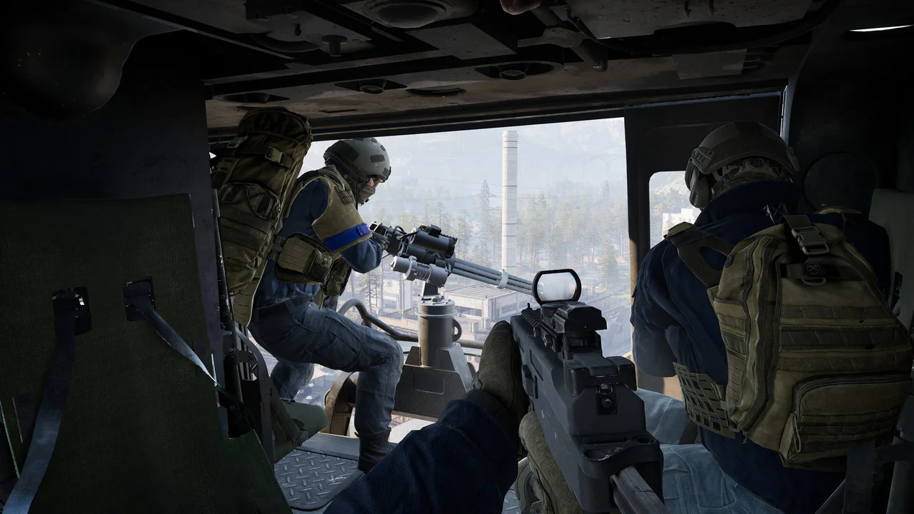 Dentro de un helicóptero de combate en Wardogs, un soldado en primer plano sostiene un rifle con mira de punto rojo mientras un compañero opera una ametralladora montada, observando una base con una torre industrial a lo lejos. Esta escena de gameplay ilustra la acción táctica y el combate vehicular presentes en el juego.